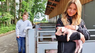 Jana und der 9-jährige Hinnerk stehen vor den Schweineställen. Jana hält ein kleines Ferkel im Arm.
