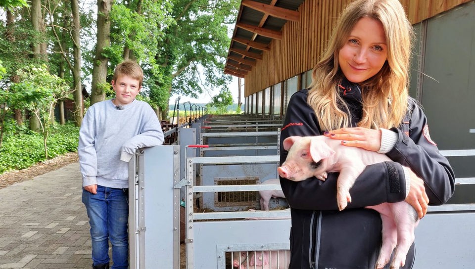 Jana und der 9-jährige Hinnerk stehen vor den Schweineställen. Jana hält ein kleines Ferkel im Arm.
