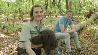 Försterin Andrea Zimmermann, Hund Cuno und Reporter Robert sitzen auf einem Baumstamm in Andreas Waldrevier.