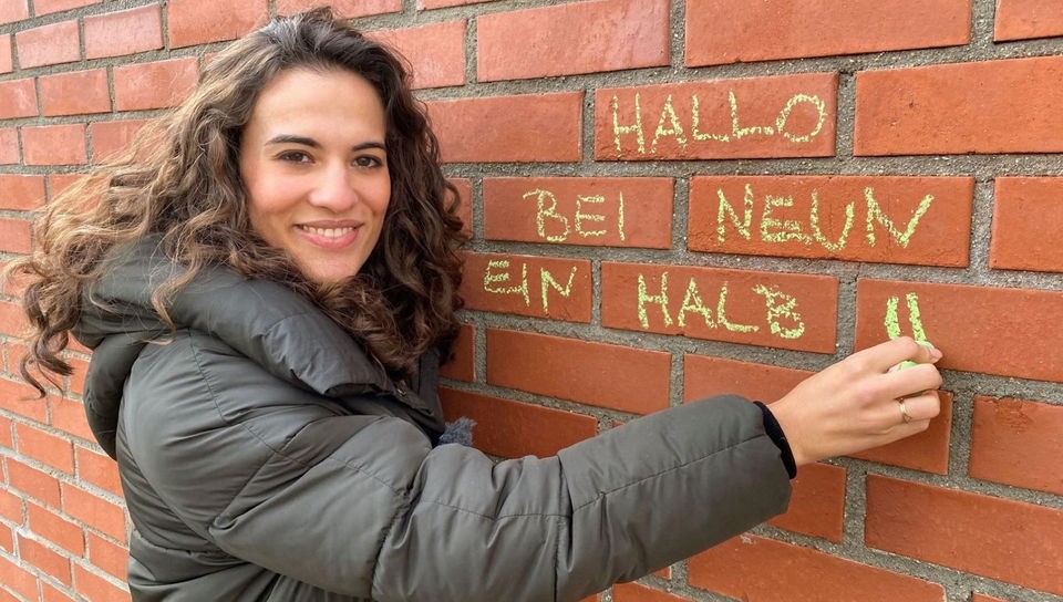 Mona schreibt mit Kreide „Hallo bei neuneinhalb“ an eine Mauer.