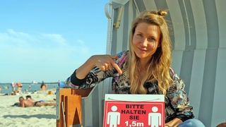Jana sitzt im Strandkorb und hält ein Schild auf dem steht, dass man 1,50 Meter Abstand halten soll.