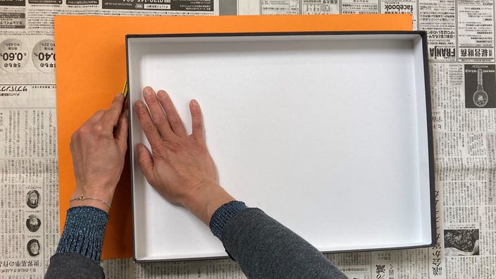 Hand malt die Kontur eines Kartondeckels auf orangener Pappe nach.