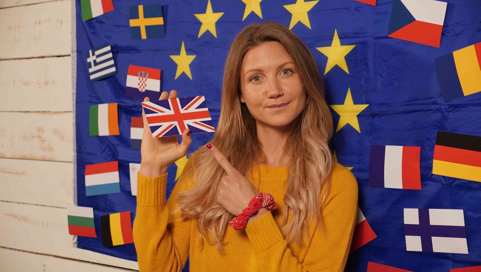 Jana vor einer weißen Holzwand mit großer EU-Flagge, in der Hand hat sie die Nationalflagge des Vereinigten Königreichs Großbritannien und Nordirland