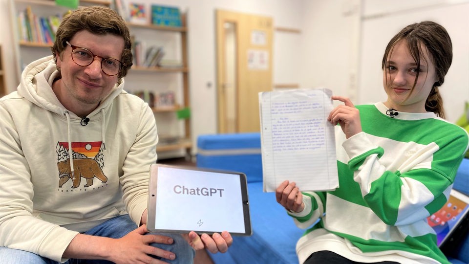 Reporter Robert und Schülerin Frida sitzen in der Schule. Robert hält ein iPad hoch, auf dem das Wort "ChatGPT" zu sehen ist. Frida hält ein Heft mit einem handgeschriebenen Aufsatz in der Hand. 