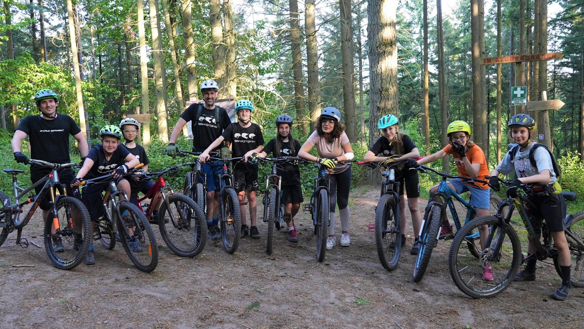 Gruppenfoto: Sieben Kinder, zwei Erwachsene und Reporterin Tessniem stehen mit Helmen und Mountainbikes im Wald und lächeln in die Kamera.