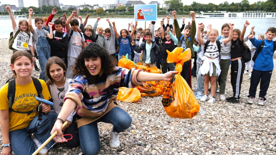 Reporterin Tessniem mit mehreren Schülerinnen und Schülern am Rhein. Vor den Kindern befindet sich ein Haufen gefüllter, orangefarbener Müllsäcke.