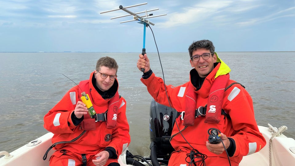 Reporter Robert und der Unterwasserlärm-Forscher Joseph „Jeff“ Schnitzler sitzen in einem Boot. Robert hält einen Sender in der Hand und Jeff eine Antenne.