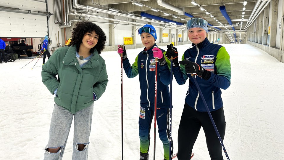 Reporterin Tessniem mit den beiden Nachwuchstalenten im Skilanglauf Magnus und Emma. Die drei stehen in der Skihalle des Thüringer Wintersportzentrums.