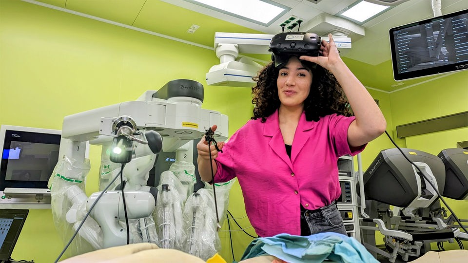 Reporterin Tessniem steht in einem OP-Saal voller Gerätschaften. Auf dem Kopf trägt sie eine Virtual Reality-Brille und hält Operationsbesteck in der Hand.