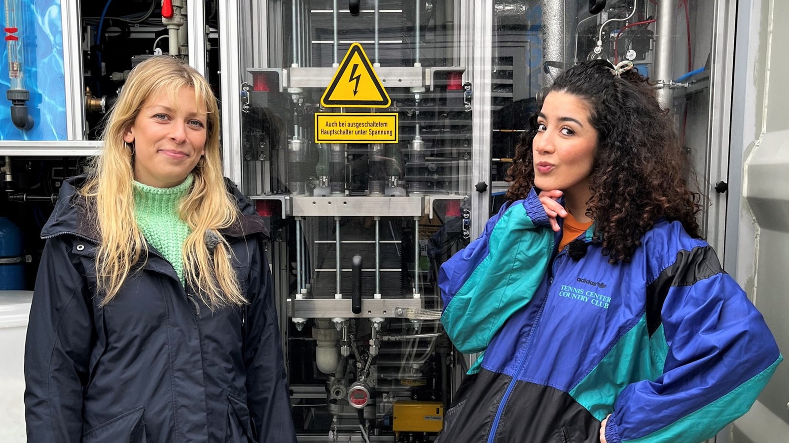 Moderatorin Tessniem steht mit der Wasserstoff-Referentin Helle Moritzen von GP Joule vor einem Elektrolyseur im Windpark in Bosbüll. Der Elektrolyseur kann Wasserstoff in die Bestandteile Wasser und Sauerstoff aufspalten.