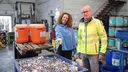 Reporterin Gesa und Florian Clever stehen vor einer großen blauen Plastikwanne voller Altbatterien in einer Recycling-Anlage. Im Hintergrund stehen bunte Plastikfässer voller Batterien. Gesa hält eine Handvoll Batterien in Richtung Kamera.