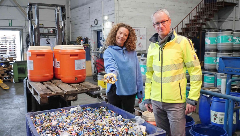 Reporterin Gesa und Florian Clever stehen vor einer großen blauen Plastikwanne voller Altbatterien in einer Recycling-Anlage. Im Hintergrund stehen bunte Plastikfässer voller Batterien. Gesa hält eine Handvoll Batterien in Richtung Kamera.