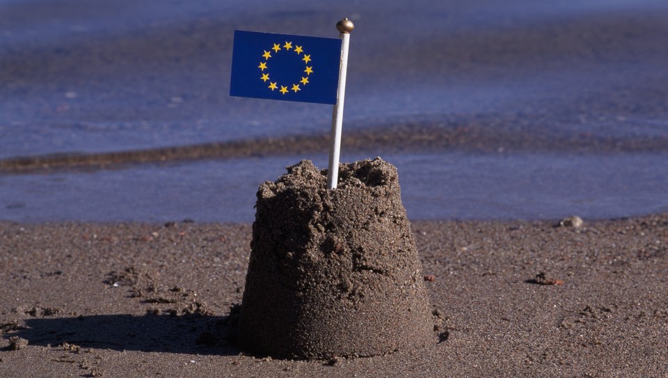 Auf einer kleinen Sandburg steckt eine EU-Flagge.