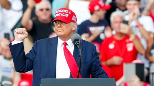 Donald Trump steht mit rotem Käppi hinter Rednerpult bei Wahlveranstaltung und reckt die Faust.