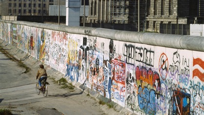 Fahrradfahrer radelt an Berliner Mauer entlang, auf der anderen Seite steht ein Wachturm.