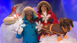 Shary und Ralph stehen mit einem Hund in einer Schnee-Kulisse. Sie tragen Anoraks, hinter ihnen liegt ein großer Schuh auf einem Eisblock.