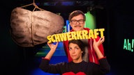 Clarissa hält das Wort "Schwerkraft" hoch, Ralph steht über ihr, hinter ihnen hängt ein großer Stein an einem Seil