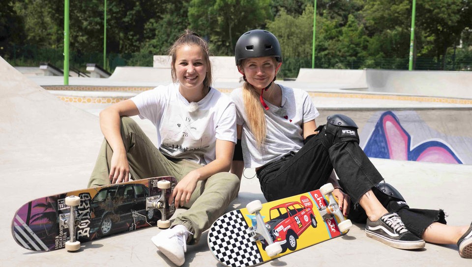 Jana und Lea sitzen mit ihren Skateboards auf einer Skateboard-Rampe. 